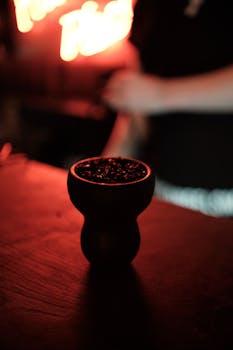 A moody, softly illuminated hookah bowl on a wooden surface in a dimly lit bar with red neon lights in the background.