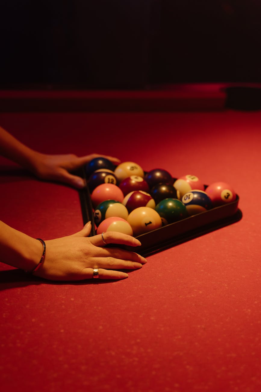 Close-up of hands arranging billiard balls on a red pool table, creating order for a game.